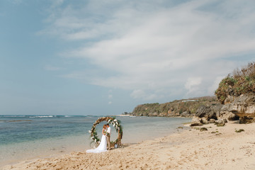 Romantick wedding cople standing on the beach near round wooden arch in boho rustc style  after ceremony by the sea. Moment of tenderness and love. Beach wedding Bali