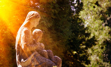 Fragment Of Ancient Stone Statue Of Virgin Mary With The Baby Jesus Christ In Sonlight Against Green Background Of Trees.