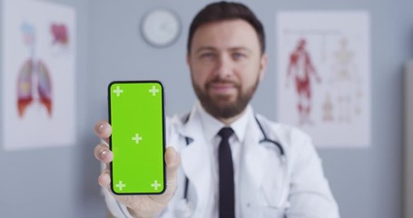 Caucasian doctor demonstrating phone with chroma key in hospital room, Portrait of handsome blue eyed man in white gown with stethoscope in medical office showing vertical smartphone with green screen