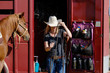 Western lifestyle shows cowgirl in cowboy hat with tack in trailer and brown horse tied.