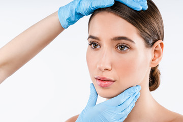 Wall Mural - Close up of plastic surgeon checking face of young woman over white background