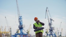 Lockdown Shot Of Middle Aged Engineer With Draw Tube Walking At Shipbuilding Yard, Talking On Cell Phone And Observing Construction Of Vessels