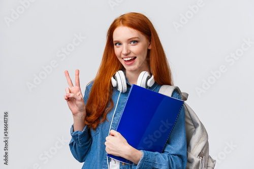 Lovely friendly college student, redhead girl with papers, documents for school, holding backpack and wear headphones over neck, make peace gesture informal greeting classmate, white background