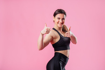 Portrait of an excited young brunette sportive muscular girl showing two thumbs up isolated over pink background. Attractive girl is happy with her figure. Ready for the summer season