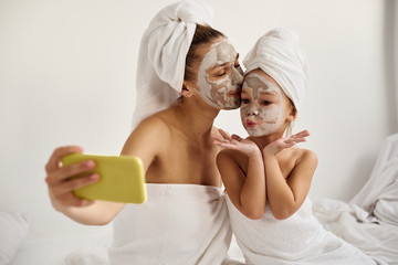 A young caucasian mother and little daughter with wrapped hair in white bath towels with a mask on their faces have a fun and make selfies on the phone