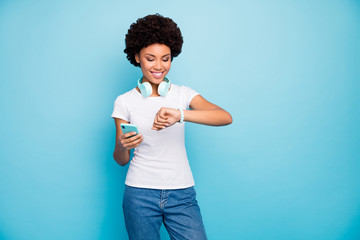 Canvas Print - Photo of pretty dark skin curly lady hipster hold telephone app hands cool modern earphones check time watches meet friends wear casual white t-shirt jeans isolated blue color background