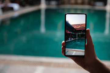 Wall Mural - hand holding phone on background of the pool in hotel.