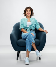 Comfort, People And Furniture Concept - Portrait Of Happy Smiling Young Woman In Turquoise Shirt And Jeans Sitting In Modern Armchair Over Grey Background
