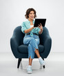 © Syda Productions - people and technology concept - portrait of smiling young woman in turquoise shirt and jeans with tablet pc computer sitting in modern armchair over grey background