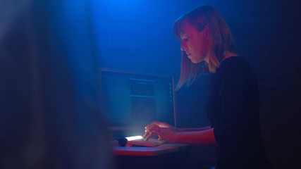Attrctive woman hacker programmer is working on a computer in a dark underground office room. Blue neon light fills the room. She enters the software to steal login information.