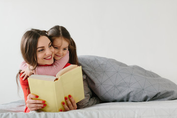 Happy family mother and child daughter reading holding book lying in bed, smiling mom baby sitter telling funny fairy tale to cute preschool kid girl having fun laughing together with bedtime stories