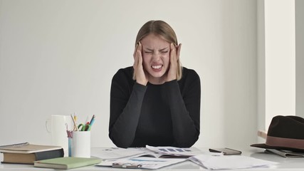 Poster - A displeased young blonde woman is feeling a headache while sitting at the table indoors in a white office