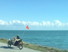 Man Riding Recumbent Bike On Road By Sea Against Blue Sky