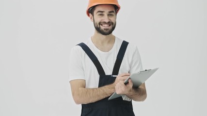 Sticker - Cheery happy young bearded man builder isolated over white wall background in helmet writing notes in clipboard.