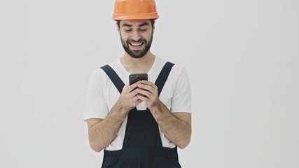 Sticker - Happy positive young bearded man builder isolated over white wall background in helmet using mobile phone