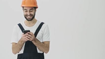 Sticker - Positive cheerful young bearded man builder isolated over white wall background in helmet using mobile phone.