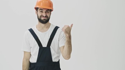 Sticker - Confused young bearded man builder isolated over white wall background in helmet pointing aside.