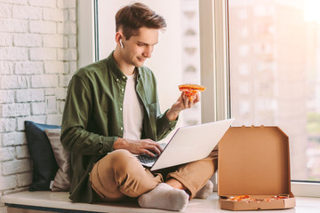 Young hipster man freelancer eating slice of tasty pizza, sit on windowsill and using laptop for study, distance learning. Businessman eat takeaway italian fast food for lunch, work remotely from home