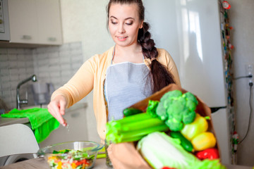 Wall Mural - Young brunette woman making healthy food on the dinner table in the kitchen. Female cooking fresh vegan salad with green vegatable