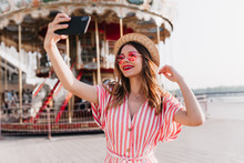 Blithesome Female Model In Striped Attire Posing Near Carousel In Straw Hat. Outdoor Shot Of Fashionable Caucasian Girl Using Smartphone For Selfie In Amusement Park.