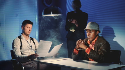 Wall Mural - Medium shot of Black prisoner in handcuffs having interview with caucasian detective in interrogation room. Female assistant making notes of the interrogation.