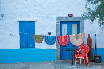 Knitting colorful cotton rag rugs, kind of carpet, are hanging on laundry thread of a Greek house with blue shutters and door. Plastic red chair and gas cylinder standing at the corner