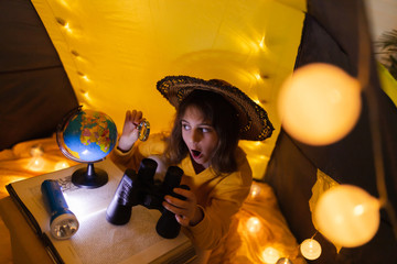 Wall Mural - young female child using magnifying glass to explore earth globe in a home made livingroom tent with
