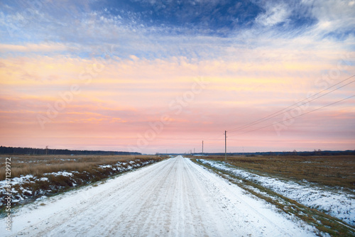 Fototapeta Pokryta śniegiem wiejska droga przez pola, zbliżenie biegunów transformatora. Las w tle. Dramatyczny zmierzchu niebo z kolorowymi chmurami. Zimowy krajobraz. Łotwa