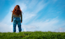 Woman With Red Hair Stands On Green Grass Looking Into The Blue Sky