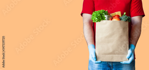 Grocery delivery courier man in red uniform and medical gloves holds paper bag with food on orange background. Safe food delivery during quarantine, online shopping or donation concept. Copy space.