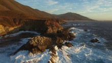 Aerial: Drone Approaching Rocky Coastline While Waves Splashing In Sea At Beach During Sunset, Scenic View Of Mountains Against Sky - Big Sur, California