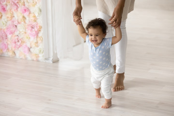 Smiling little biracial toddler infant child make first steps at home holding mom hands, happy small african American baby learn walking with mother support and care, upbringing, childcare concept