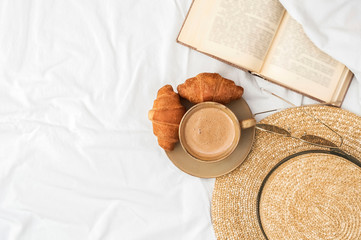 Coffee cup with foam, croissants, summer hat, reading glasses and open book on a white bedsheet. Background with space. Delicious breakfast . Top view.  Flat lay