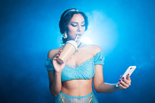 Magic, Cosplay And Fairy Tale Concept - Portrait Of A Young Woman In The Image Of An Eastern Fairy Princess Holds Smartphone And Shaker On Blue Background.
