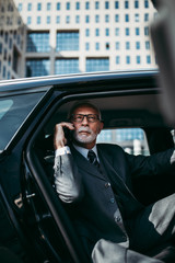 Wall Mural - Good looking senior business man sitting on backseat in luxury car. He opens car doors and going or stepping out. Big business building in background. Transportation in corporate business concept.