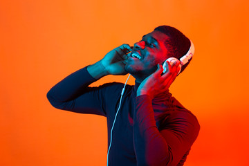 Wall Mural - Young african-american man's listening to music in headphones in neon light. Male portrait. Concept of human emotions, facial expression, holidays or weekend, hobby,