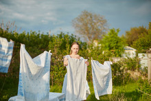 Blonde Woman Villager Hanging Wet White-blue Laundry On Clothesline To Dry In The Backyard. A Green Tree In The Background.