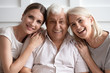 © fizkes - Portrait of happy adult family sit rest on couch smile for picture at home together, overjoyed senior parents hug embrace grownup daughter relax enjoy leisure weekend, look at camera posing