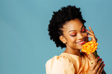Wall Mural - Portrait of a happy young woman holding yellow orange rose and smiling with eyes closed, isolated on blue background