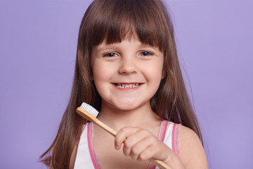 Wall Mural - Close up portrait of cheerful positive kid looking directly at camera, smiling sincerely, brushing teeth, being in high spirits, standing isolated over lilac background in studio. Childhood concept.