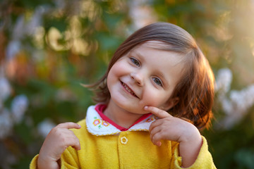 Happy smiling little girl wearing yellow jacket, looking directly at camera, touching her cheek with fore finger, female kid with dark hair and charming smile. Childhood, children emotions concept.