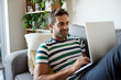 © mavoimages - Smiling young man relaxing on his sofa using a laptop