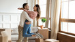 Happy young couple celebrating moving day, hugging in new apartment, loving husband lifting smiling wife, standing in living room with cardboard boxes with belongings, relocation and mortgage