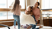 Happy Adorable Little Daughters With Young Mother Pillow Fighting In Bedroom, Laughing And Jumping, Two Cute Preschool Girls Having Fun With Mum, Playing Active Funny Game At Home