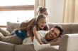 © fizkes - Two happy little girls playing lying on couch with father, smiling young dad having fun with preschool daughters, laughing, relaxing on cozy sofa at home, funny family activity, enjoying weekend