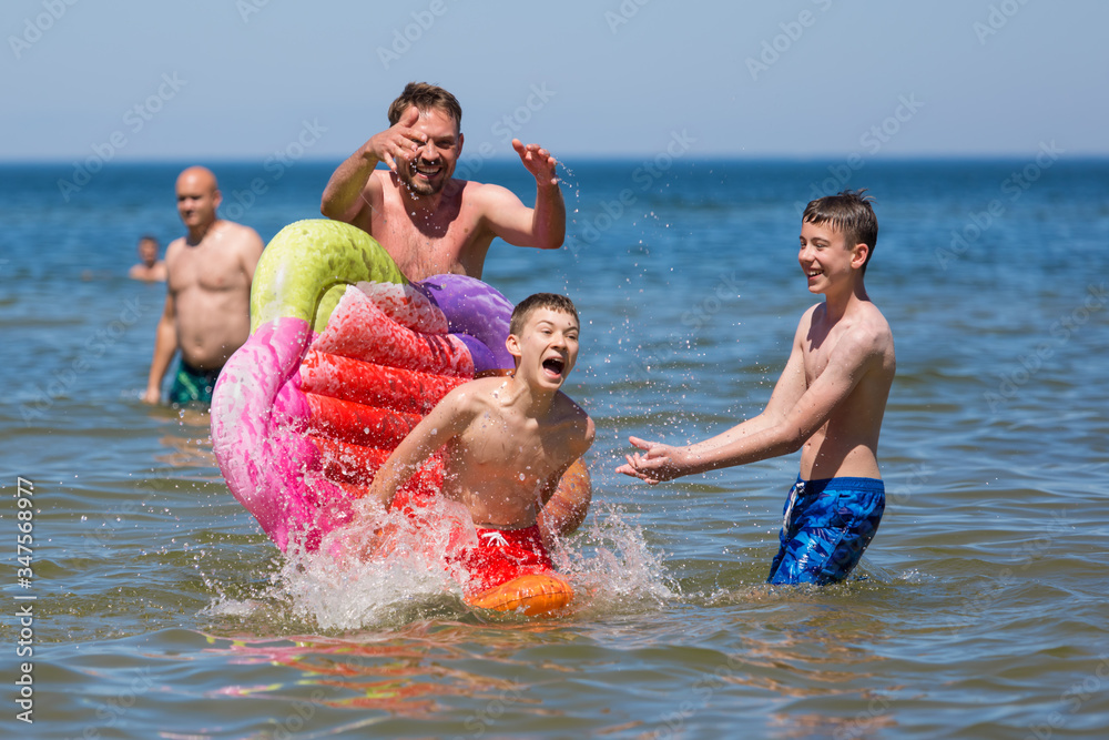 Zabawa w morzu, roześmiana rodzina chlapie się wodą, chłopiec spada z kolorowego materaca - obrazy, fototapety, plakaty 