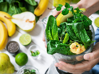 Woman is preparing a healthy detox drink in a blender - a smoothie with fresh fruits, green spinach and superfood seeds. Healthy lifestyle concept, ingredients for smoothies on the table, top view