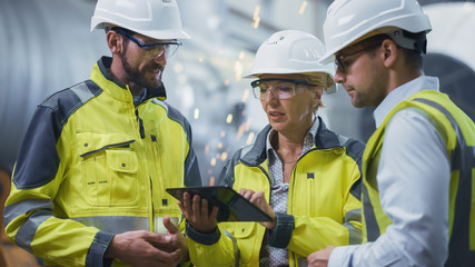 Wall Mural - Three Heavy Industry Engineers Stand in Pipe Manufacturing Factory, Use Digital Tablet Computer, Have Discussion. Large Pipe Assembled. Design and Construction of Oil, Gas and Fuels Transport Pipeline