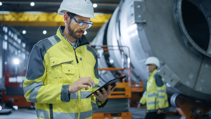Wall Mural - Heavy Industry Engineer Stands in Pipe Manufacturing Factory, Use Digital Tablet Computer. Facility for Construction of Oil, Gas and Fuel Pipeline Transportation Products.