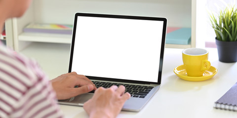 Back shot of smart man typing on white blank screen computer laptop that putting on white working desk and surrounded by coffee cup, potted plant and stationary.